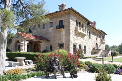 A large stone house with a red-tiled roof, surrounded by gardens and sculptures of two seated figures.