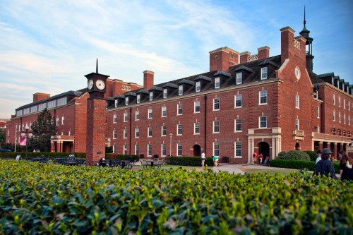 A brick building with a clock tower, surrounded by greenery, under a blue sky with scattered clouds.