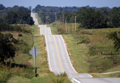 A winding road stretches through green fields under a clear blue sky, with trees lining the sides.