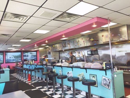 A retro diner interior with pink accents, checkered floor, and a cook at the grill behind the counter.