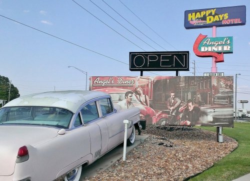 A vintage car parked near a retro diner sign, featuring "Angel's Diner" and "Happy Days Hotel," with a clear blue sky.