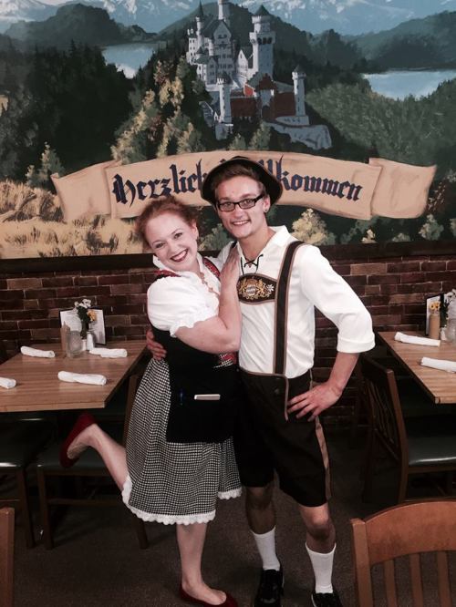 A woman in traditional German attire poses with a man in lederhosen, smiling in a restaurant with a scenic mural.