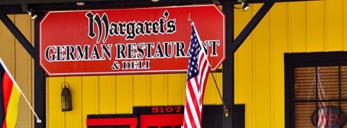 Sign for "Margaret's German Restaurant & Deli" with an American flag and colorful building background.