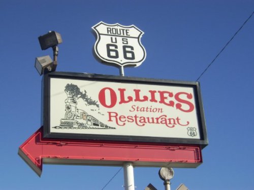 Sign for Ollie's Station Restaurant on Route 66, featuring a train graphic and an arrow pointing left. Clear blue sky background.