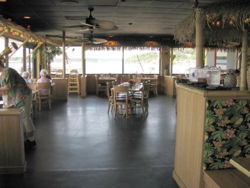 A spacious restaurant interior with wooden tables, chairs, and tropical decor, featuring large windows and a view outside.