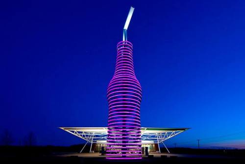 A tall, illuminated structure resembling a bottle, featuring purple and white stripes against a twilight sky.
