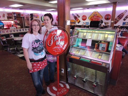 Two waitresses stand beside a vintage jukebox in a retro diner, with colorful decor and signage in the background.