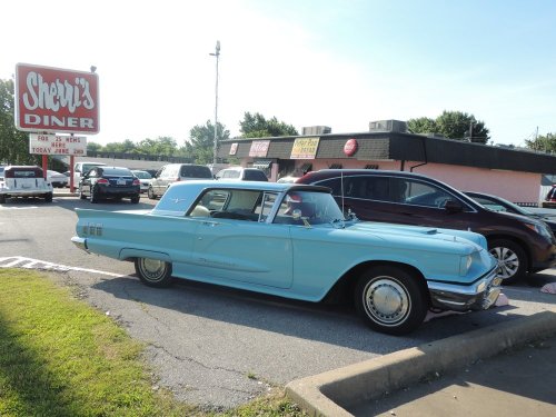 A vintage blue car parked outside Sherri's Diner, with modern vehicles in the background and a clear sky.
