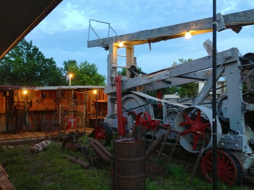 An old, rustic machinery setup with red wheels, surrounded by trees and a cloudy sky at dusk.
