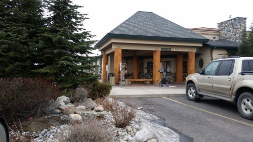 A building with a sloped roof and wooden pillars, surrounded by shrubs and rocks, with a parked SUV in the foreground.