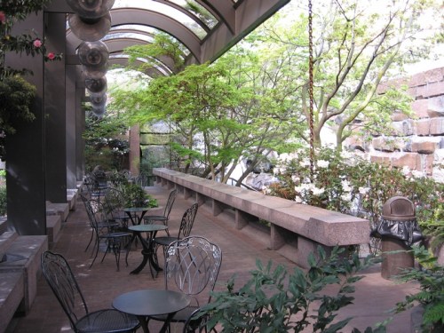 A serene outdoor seating area with metal tables and chairs, surrounded by greenery and a stone wall.