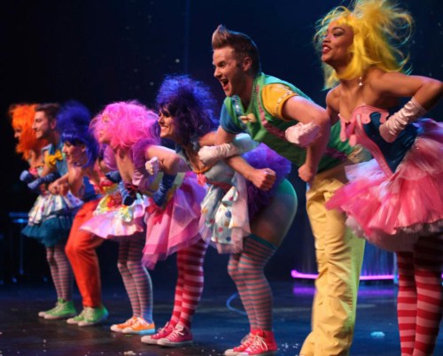 A colorful group of performers in vibrant costumes, striking playful poses on stage with bright wigs and cheerful expressions.
