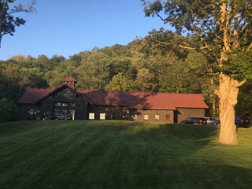 A large, dark wooden building with a red roof, surrounded by green trees and a manicured lawn under a clear blue sky.