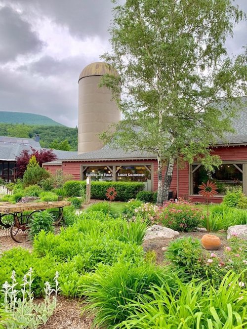 A rustic building with a silo, surrounded by lush greenery and colorful flowers under a cloudy sky.