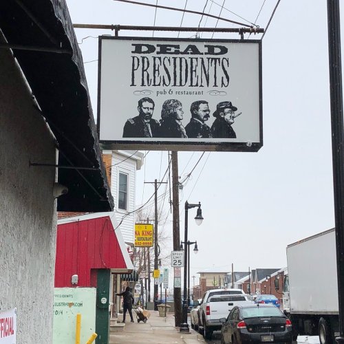 Sign for "Dead Presidents" pub and restaurant featuring four stylized figures, set against a snowy street scene.