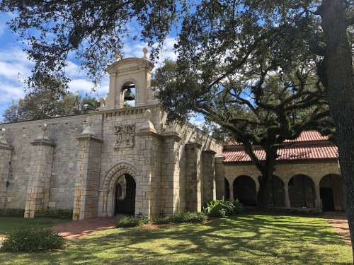 Historic stone building with a bell tower, surrounded by lush greenery and trees under a partly cloudy sky.