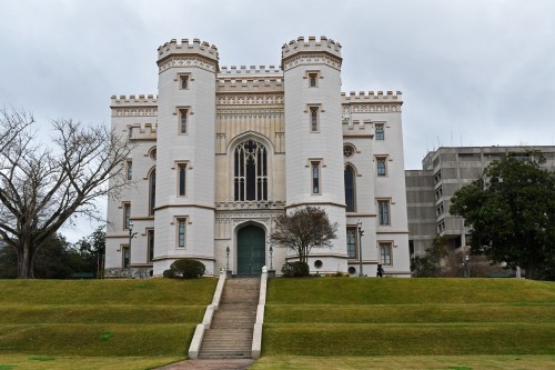 A historic castle-like building with towers, large windows, and a manicured lawn in front, set against a cloudy sky.