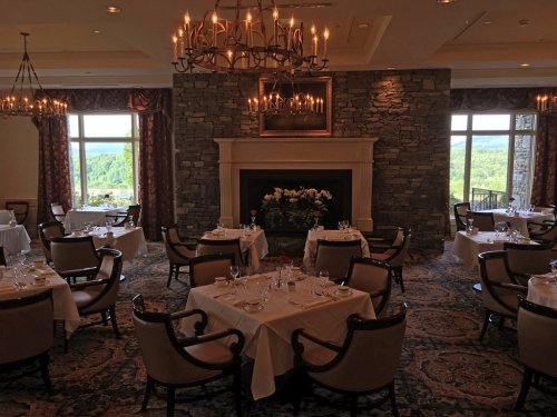 Elegant restaurant interior with tables set for dining, a stone fireplace, and large windows showcasing a scenic view.