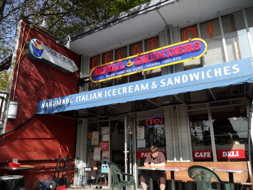 Exterior of a deli with a sign for "Rola's Grinders," featuring gelato and sandwiches, with outdoor seating.