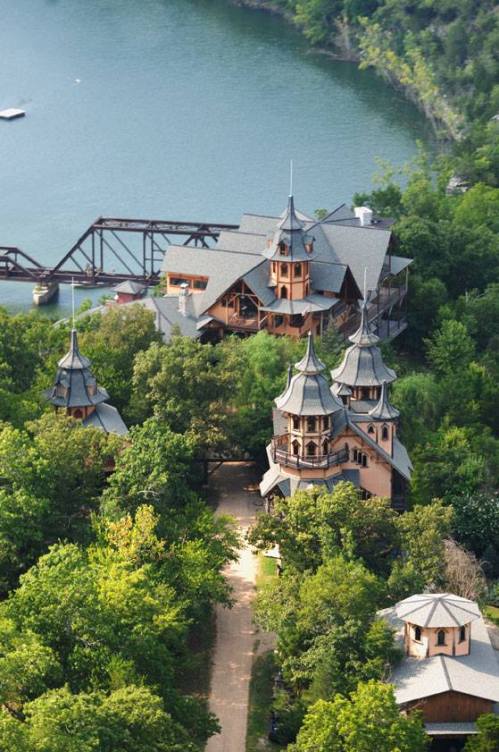 Aerial view of unique, multi-turreted buildings surrounded by lush greenery near a calm body of water.