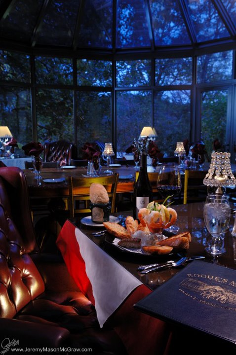 A cozy dining setting with elegant decor, featuring a table with gourmet food, wine, and a menu in a glass-enclosed space.
