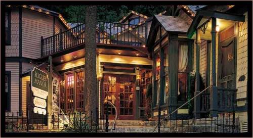 A charming, illuminated Victorian-style house with a sign reading "Piggin Manor," surrounded by trees and a wrought-iron fence.