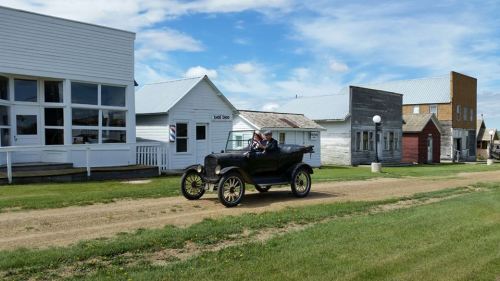 A vintage car drives down a dirt road past historic buildings under a blue sky with scattered clouds.