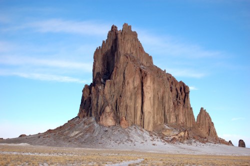 A towering rock formation rises against a clear blue sky, surrounded by a barren landscape.