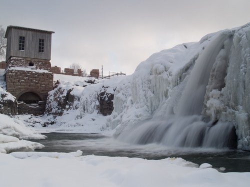 A frozen waterfall cascades over icy rocks, with a historic building nearby, surrounded by snow and a cloudy sky.