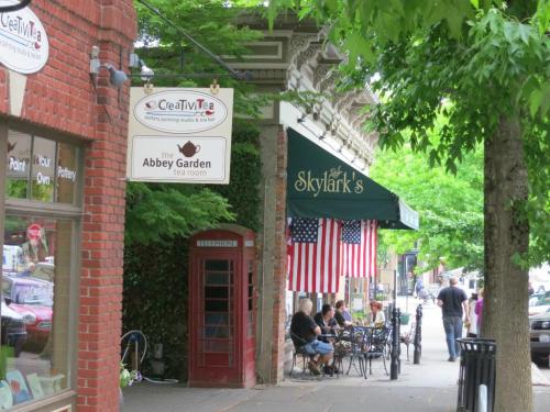 A street scene featuring a red phone booth, shops, and outdoor seating with American flags and greenery.