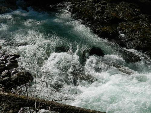 Rushing water flows over rocks in a stream, creating white frothy waves and a vibrant turquoise hue.
