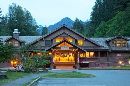 A rustic lodge surrounded by trees, with warm lights glowing from the windows and mountains in the background.