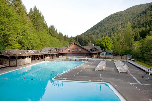 A serene outdoor pool area surrounded by trees and mountains, featuring lounge chairs and a rustic lodge in the background.