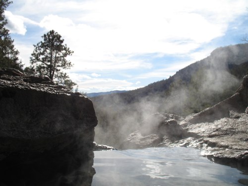 A serene hot spring surrounded by rocky terrain and mist, with mountains and a cloudy sky in the background.