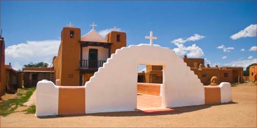 A white archway leads to a historic adobe building with crosses, set against a blue sky with fluffy clouds.