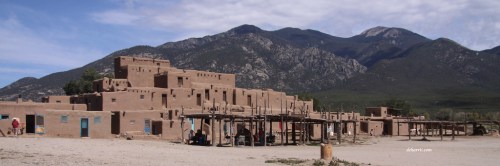 A traditional adobe pueblo structure against a backdrop of mountains and a clear sky.