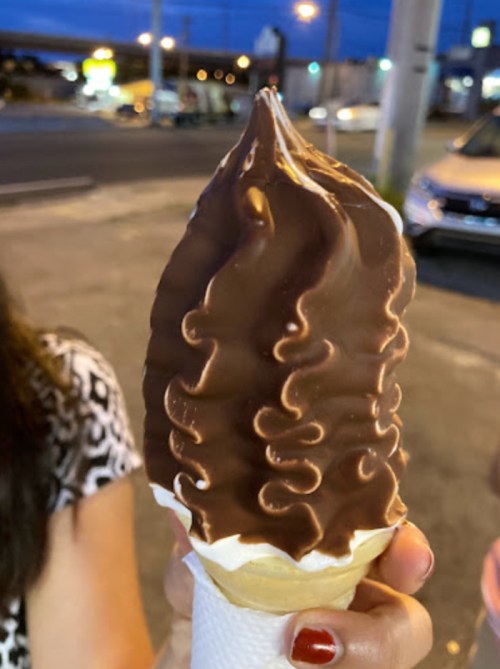 A hand holding a chocolate-dipped soft serve ice cream cone with a swirl of vanilla on a street at dusk.