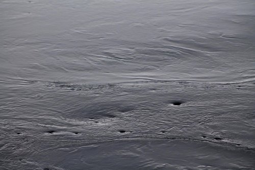 A close-up of dark, rippling water with subtle waves and small circular patterns on the surface.