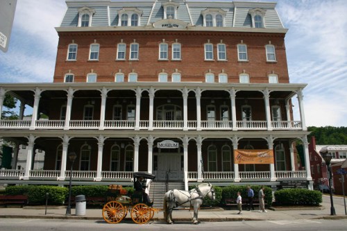 Historic building with a large porch, featuring a horse-drawn carriage in front and people walking nearby.