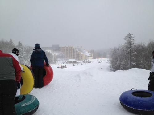 People standing on a snowy hill with colorful snow tubes, overlooking a ski resort in the distance.
