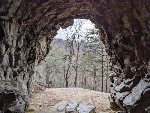 View from inside a rocky cave, framing a forested landscape with trees and a cloudy sky in the background.