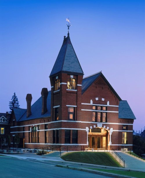 Historic brick building with a pointed roof and tower, illuminated at dusk against a blue sky.