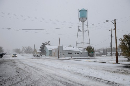 A snowy street scene featuring a water tower, buildings, and a car driving through a winter storm.