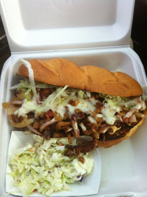 A large sandwich filled with meat, lettuce, and sauce, served with a side of coleslaw in a takeout container.