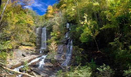 A scenic view of two waterfalls surrounded by vibrant autumn foliage and rocky terrain under a blue sky.