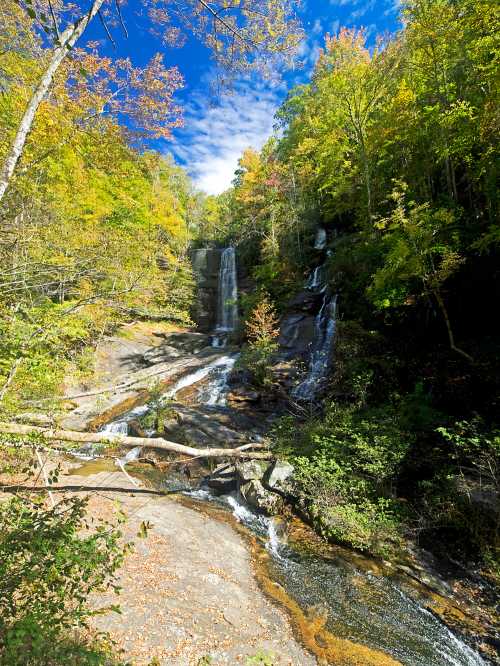 A serene waterfall cascades down rocky cliffs, surrounded by vibrant autumn foliage and a clear blue sky.