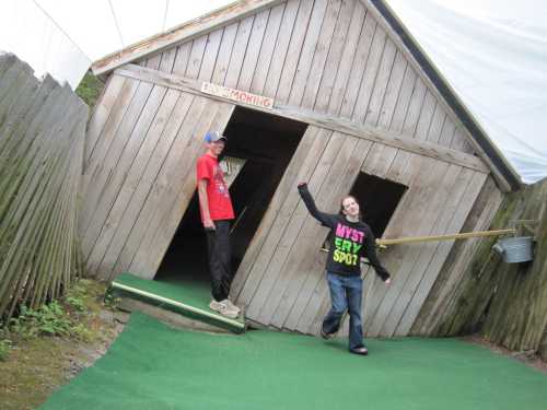 Two children playfully pose outside a tilted wooden house with a "No Smoking" sign, surrounded by greenery.