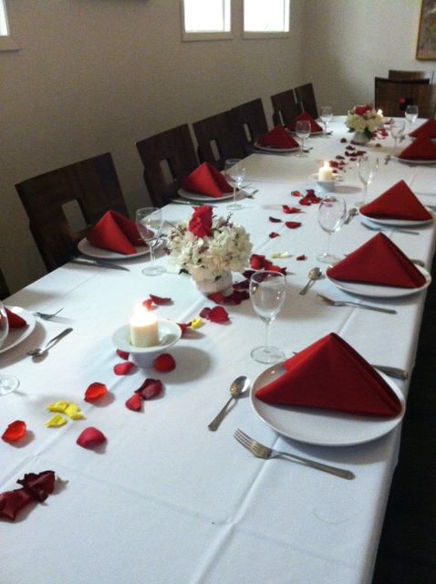 A beautifully set dining table with red napkins, rose petals, candles, and floral centerpieces.