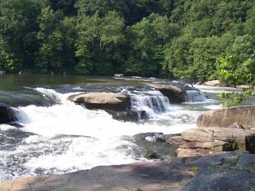 A serene river scene with cascading waterfalls over large rocks, surrounded by lush green trees.