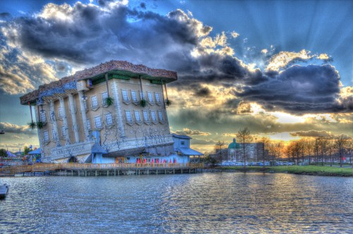 A tilted, whimsical building with a grassy roof, set against a dramatic sky and reflecting in the water below.